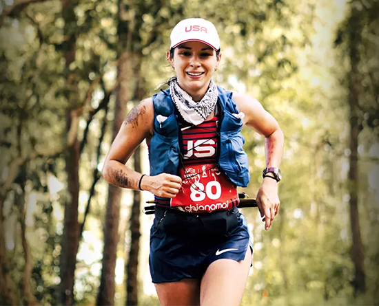 A woman in a white ball cap and a red running jersey jogs through the woods.
