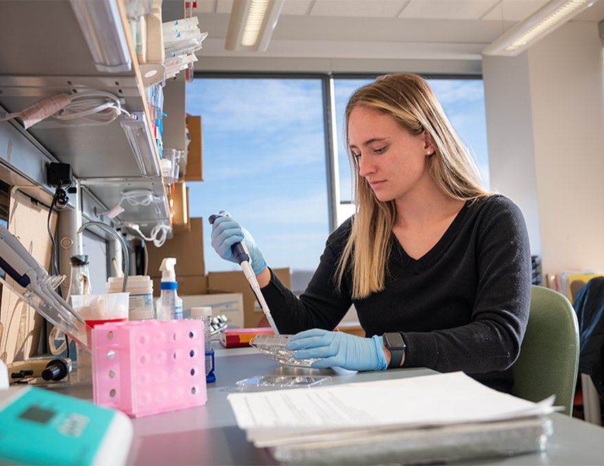 A woman in a black shirt uses a pipette at a lab desk.