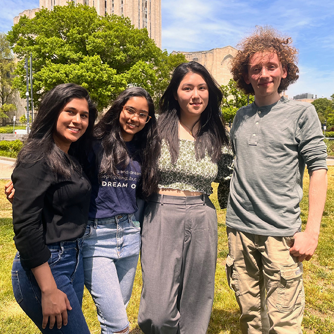 Four students stand with their arms around each other in a grassy plaza.