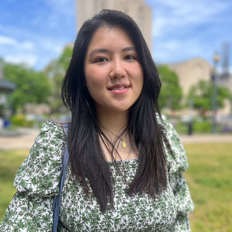 A young woman in a green-and-white shirt stands in a grassy plaza.