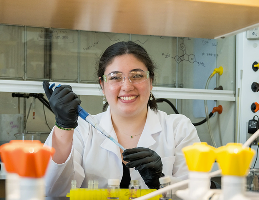 A young woman in a lab coat smiles while holding a syringe.