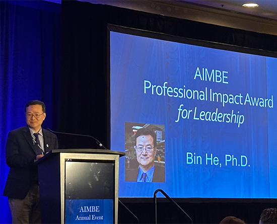 A man in a black suit stands at a podium while a screen to the right says, "A.I.M.B.E. Professional Impact Award for Leadership. Bin He, Ph.D."