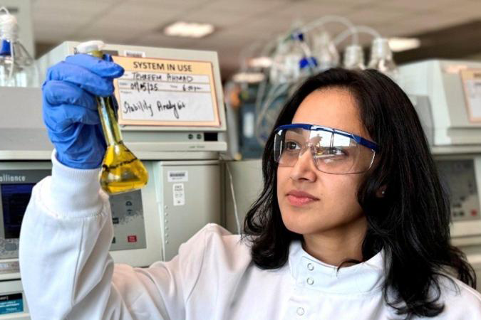 student works in lab, looks at beaker filled with yellow liquid